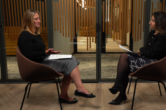Two women are seated in a glass-walled room, engaged in a conversation during a formal interview or discussion, with both holding papers on their laps.