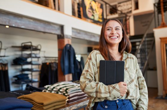 A smiling young woman with long brown hair holding a tablet, standing in a clothing store with folded sweaters on a table behind her.