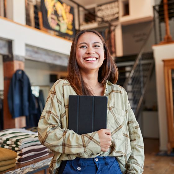 A smiling young woman with long brown hair holding a tablet, standing in a clothing store with folded sweaters on a table behind her.
