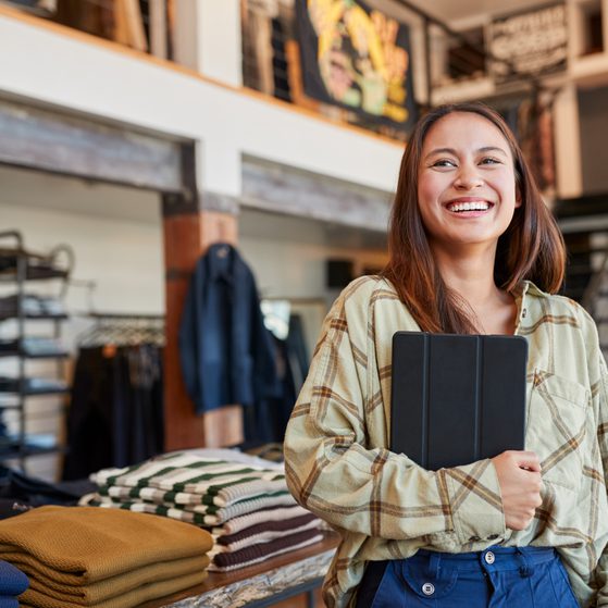 A smiling young woman with long brown hair holding a tablet, standing in a clothing store with folded sweaters on a table behind her.