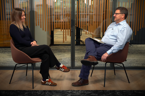 A woman and a man are sitting across from each other in a glass-walled office, engaged in conversation. The woman has long blonde hair, is dressed in a dark sweater and trousers, and is wearing distinctive leopard-print sneakers. The man has short dark hair, glasses, a light striped shirt, and dark trousers, with a notebook on his lap. They are seated in brown chairs in front of a glass wall with wooden vertical stripes.