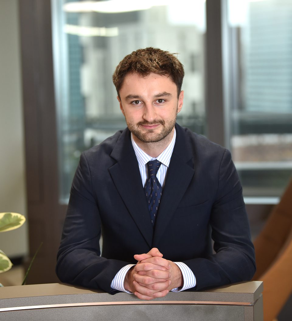 A man in a dark suit with a striped shirt and tie, sitting at a table with hands clasped, in an office with large windows and modern furniture.