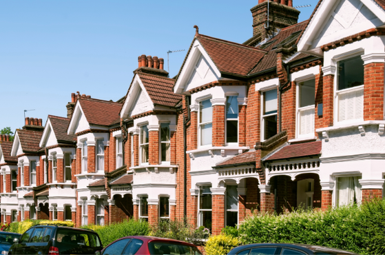 Row of red-brick Victorian-style terraced houses with white detailing, pitched roofs and chimneys, along a sunny street with parked cars and greenery.