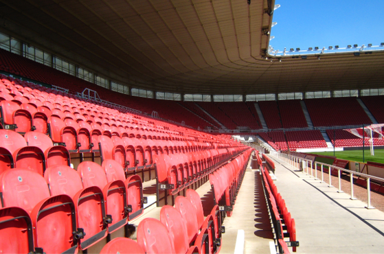 Empty red stadium seats under a partly cloudy sky, with a view of the football pitch and goalposts in the background.