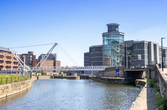 A riverside urban scene with a calm canal, a modern glass tower, a white cable-stayed pedestrian bridge, brick warehouses on the left, and a stone quay on the right.