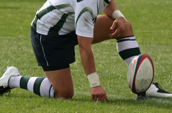 A rugby player kneeling on the grass with one knee and one hand on the ground, holding a rugby ball in front of his foot.