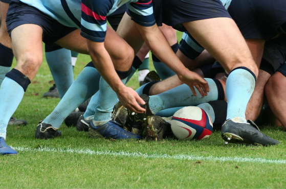 Rugby players in a scrum, with some players crouching and pushing, focusing on the ball on the ground, on a grassy field.