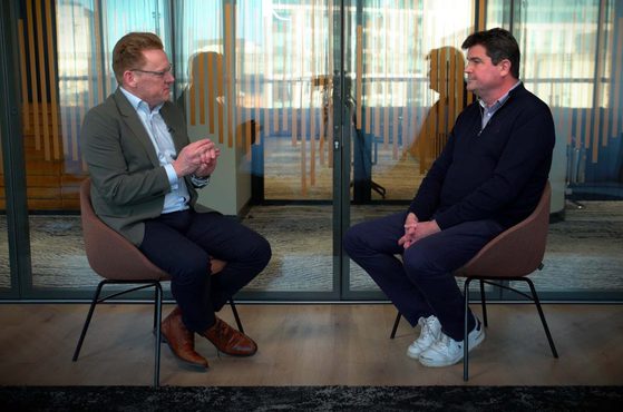 Two men are seated facing each other on chairs in a room with a glass partition behind them. They appear to be engaged in a conversation.