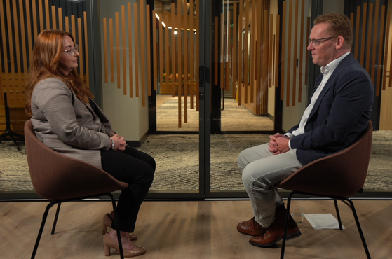 A woman and a man sit facing each other in a room with glass doors and wooden paneling, engaged in conversation. The woman has long red hair and glasses, the man wears glasses and a navy blazer.