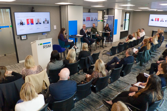 Panel discussion in a bright conference room, with an RSM-branded podium, panelists at a high table, and an audience watching large screens.