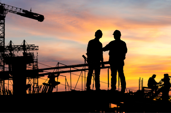 Silhouetted construction site at sunset with two workers on a platform facing each other, a crane and scaffolding outline against an orange-pink sky.