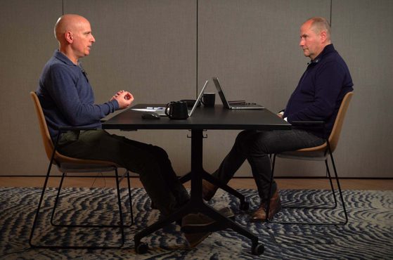 Two men sit facing each other at a black table in a room with grey walls. The man on the left is bald, wearing a blue sweater, and has his hands clasped. The man on the right has short hair, is dressed in dark clothing, and has his hands resting on his lap. There are two laptops, a black mug, and some papers on the table.