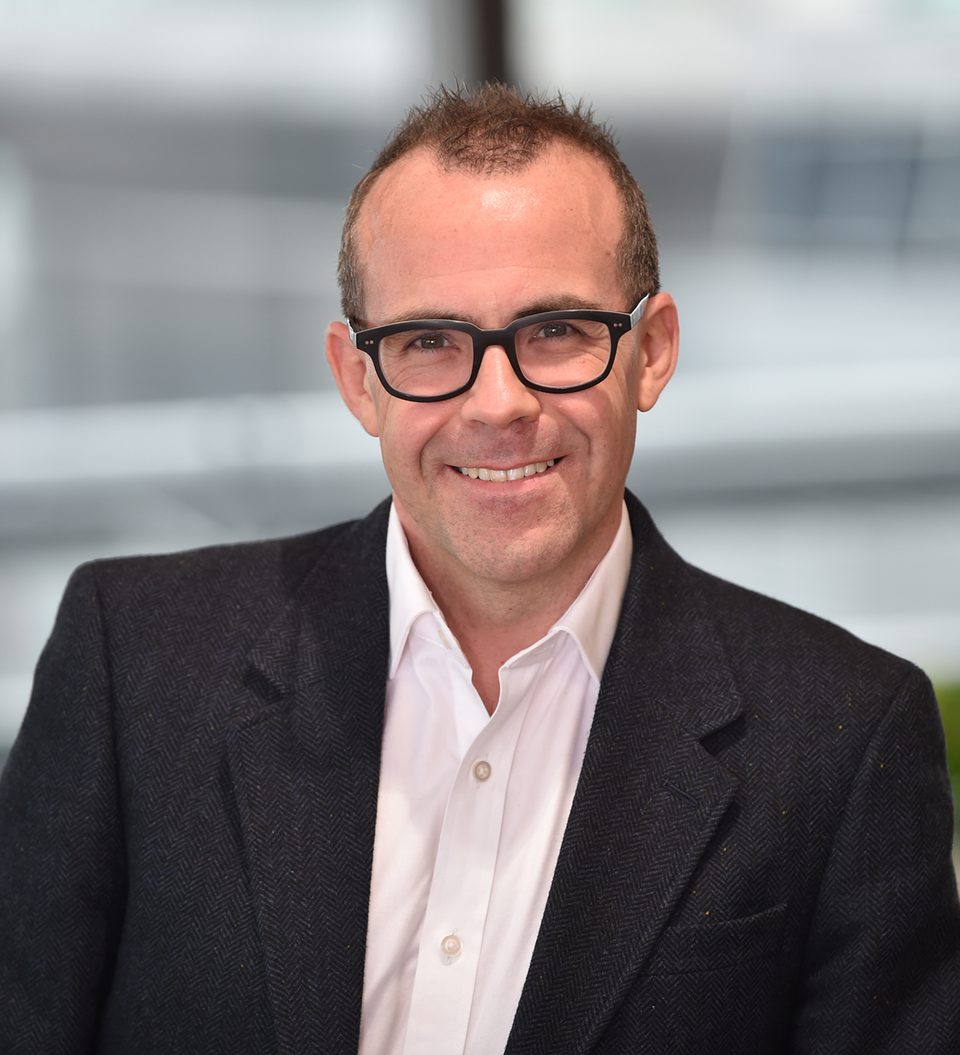 A man with short brown hair, wearing black glasses, a white shirt, and a dark blazer, smiling in a modern indoor setting with blurred architectural background.