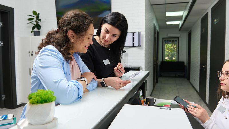 Two women are at a reception desk, with one showing a document to the other. A third woman is seated nearby holding a smartphone, in a modern office setting.