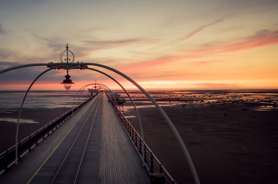 A long pier extends into the ocean at sunset, with soft pastel colours in the sky, and gentle waves beneath it. The pier has modern arched supports and lighting.