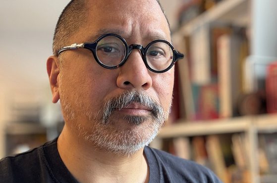 Person with round black glasses, short dark hair and a grey beard, wearing a black T-shirt, in a room with blurred bookshelves in the background