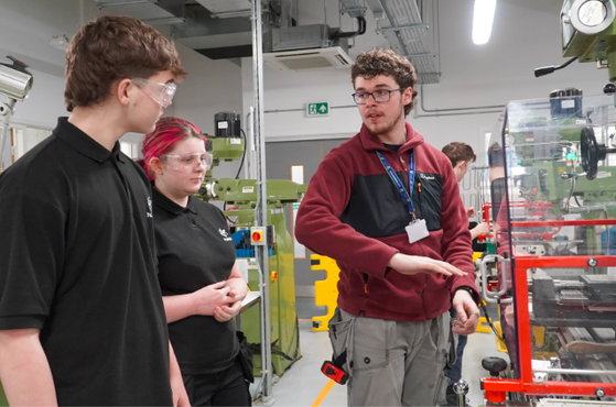 Three students wearing safety goggles observe a technician explaining machinery in a workshop with green equipment and safety barriers.