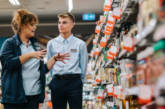 Two store employees, one with curly hair and the other with short hair, converse in an aisle lined with shelves of various products.