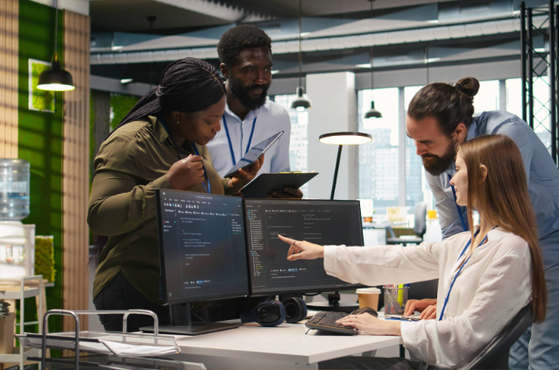 Four colleagues collaborate around a desk with dual computer monitors in a modern office, with large windows and bright lighting.