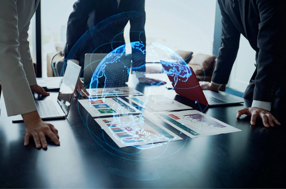 People in business suits leaning over a table with laptops and documents, with a digital hologram of a globe and data visualisation projecting above.