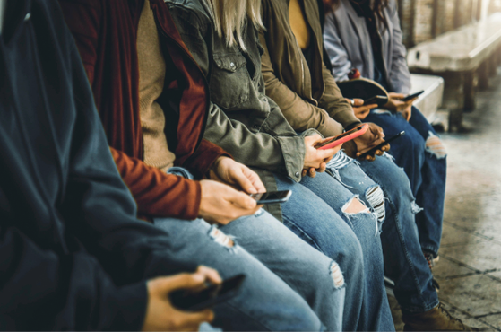 People seated on a bench beside a platform, all absorbed in their smartphones, wearing casual jackets and ripped jeans.