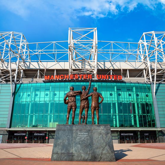 Bronze statue of three football players with arms raised in front of Manchester United stadium, glass frontage and white steel framework against a blue sky