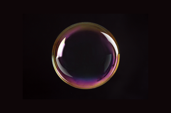 A single iridescent soap bubble floats against a dark background, its glossy surface reflecting purple and pink hues with a thin gold rim and bright highlights.