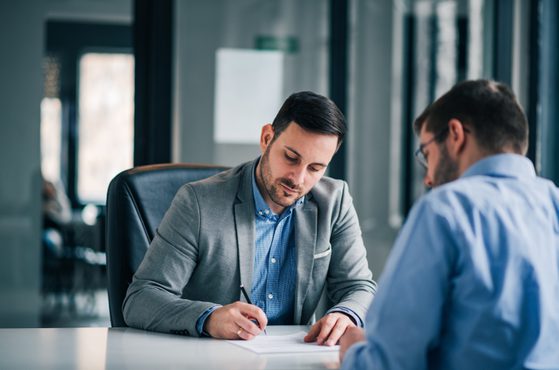 Two men in business attire sit at a table in an office, engaged in a serious discussion, with documents and a pen on the table.