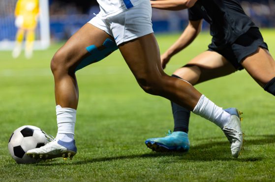 Two football players contesting the ball on a green pitch, one in white shorts and socks, the other in dark kit; a black-and-white football beside their feet.