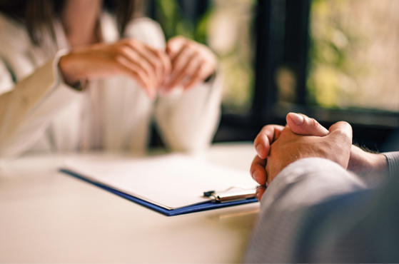 Two people sit at a bright table with a clipboard and papers; one clasps their hands while the other gestures in a thoughtful discussion.