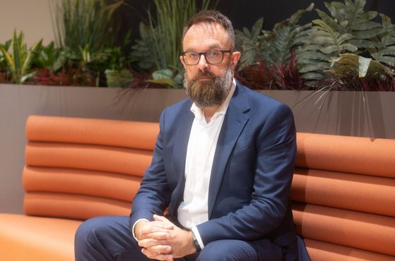 A bearded person wearing glasses and a navy suit with a white shirt sits on an orange cushioned bench, beside a planter with green foliage.