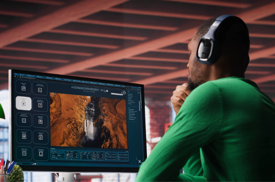 A man wearing headphones, sitting at a desk in an industrial-style room, working on a computer with an aerial landscape image on the screen.