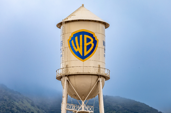 A beige water tower with the Warner Bros logo in blue and yellow, set against a cloudy sky and distant mountains.