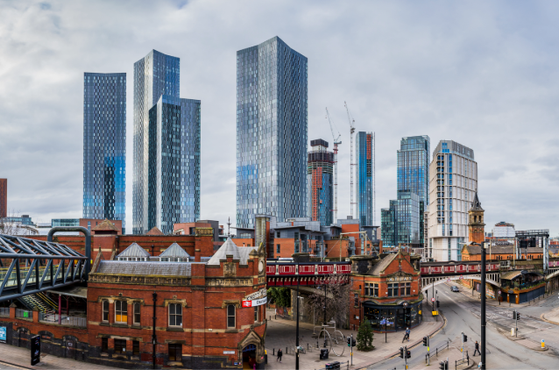 Panoramic cityscape showing a riverfront with red-brick Victorian buildings in the foreground and tall glass skyscrapers rising behind them under a cloudy sky.