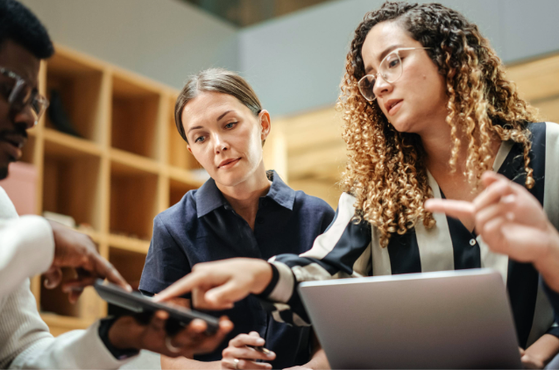Four people in a discussion, with one person pointing at a tablet, in a room with wooden shelves and a neutral background.