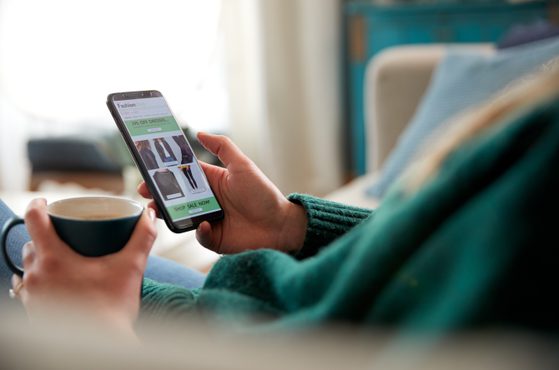 A person sitting on a sofa, holding a smartphone displaying an online fashion sale, with a cup of coffee in their other hand, in a cozy living room.
