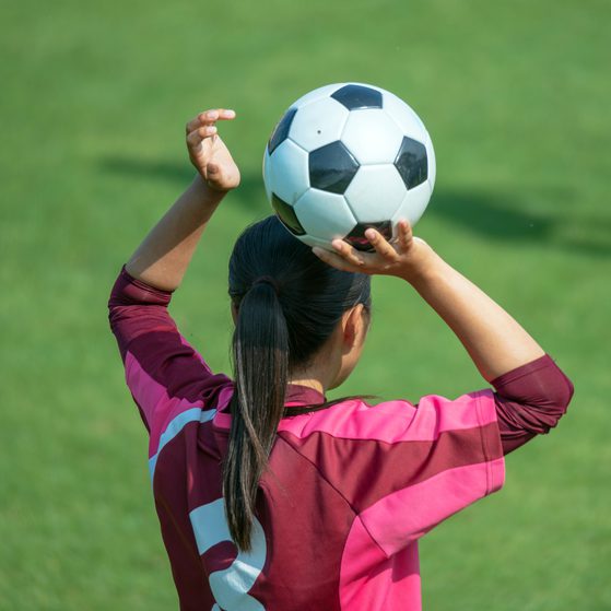A girl with a ponytail, wearing a pink and maroon sports jersey, prepares to throw a soccer ball during a game on a grassy field.