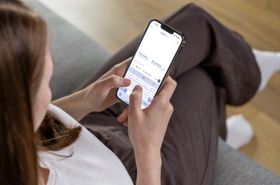 A person sitting on a grey sofa using a smartphone with a white screen and visible keyboard, wearing brown trousers and white socks.
