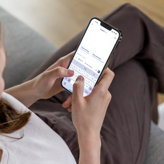 A person sitting on a grey sofa using a smartphone with a white screen and visible keyboard, wearing brown trousers and white socks.
