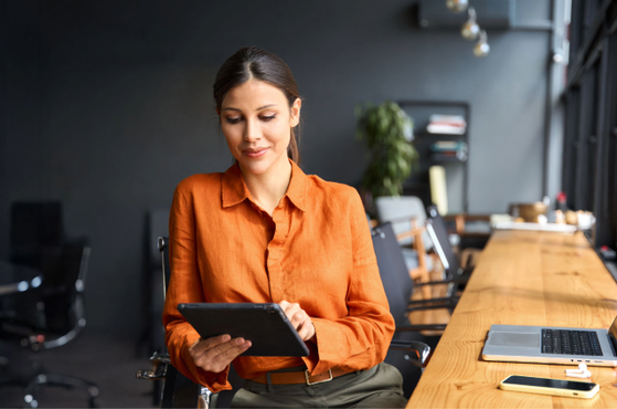 A person in an orange shirt sits at a long wooden desk in a bright coworking space by large windows, looking at a tablet with a laptop nearby.