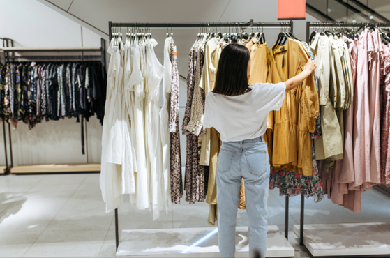 Person with shoulder-length dark hair in a white top and light jeans browses dresses on racks in a boutique, examining white, beige and pink garments.