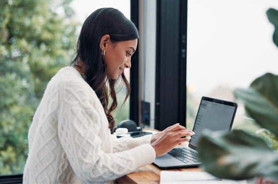 A woman in a white cable-knit sweater sitting at a wooden desk near a window, working on a laptop with green foliage outside.