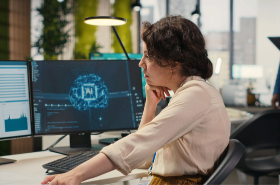 Person seated at a desk with two monitors displaying charts and a neural network graphic, resting their chin on a hand in a bright office with colleagues in the background.
