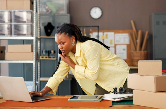 A woman in a yellow shirt looking at her laptop screen in a storage room or warehouse, with packing boxes and shelves in the background.