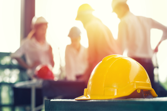 Bright yellow hard hat rests on a dark surface in the foreground, with a blurred group of construction workers wearing helmets in sunlight in the background.