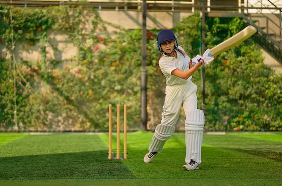 A girl in a cricket uniform, wearing a helmet and pads, is preparing to hit a cricket ball with a bat on a grassy field with trees in the background.