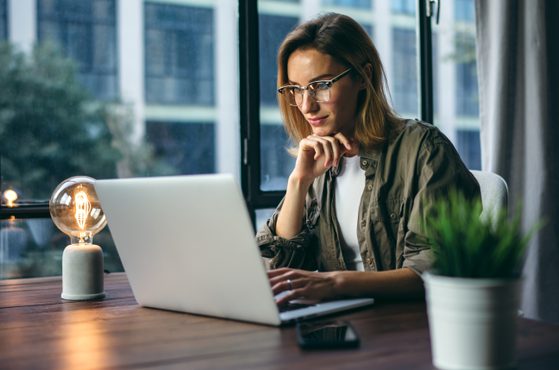 A woman with glasses sits at a desk, working on a laptop, with her chin resting on her hand, in a modern office with large windows and a plant on the desk.