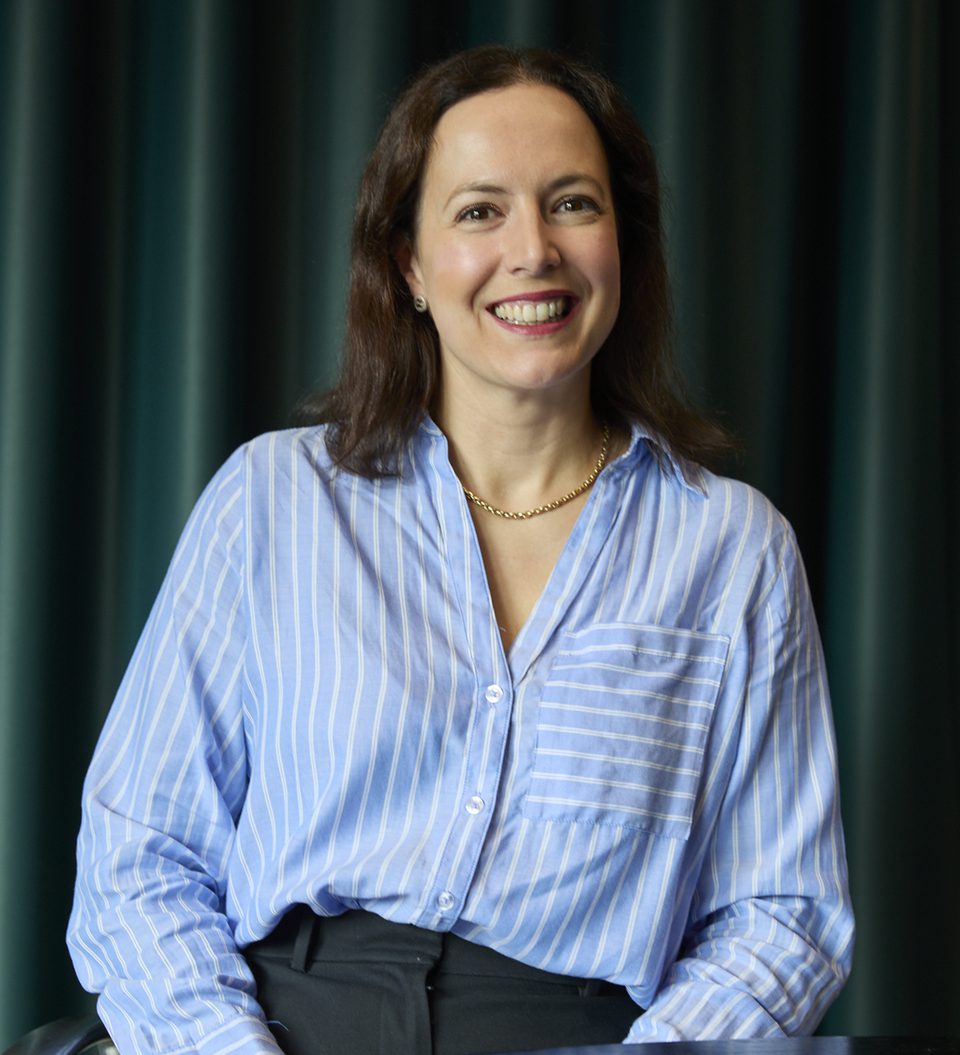 Smiling person with brown hair wearing a light blue striped shirt and a gold necklace, seated at a table with dark curtains in the background.