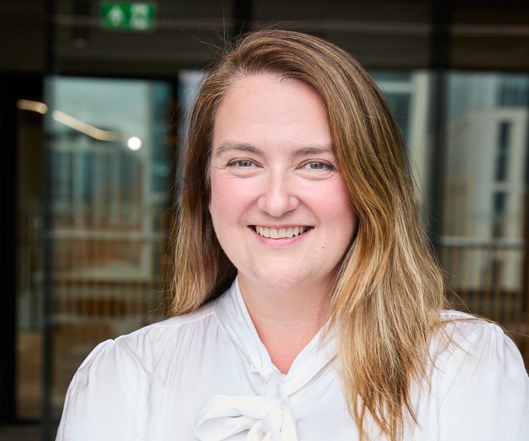 Portrait of a person with long light brown hair, wearing a white blouse with a bow, smiling with arms crossed in a bright office lobby backdrop