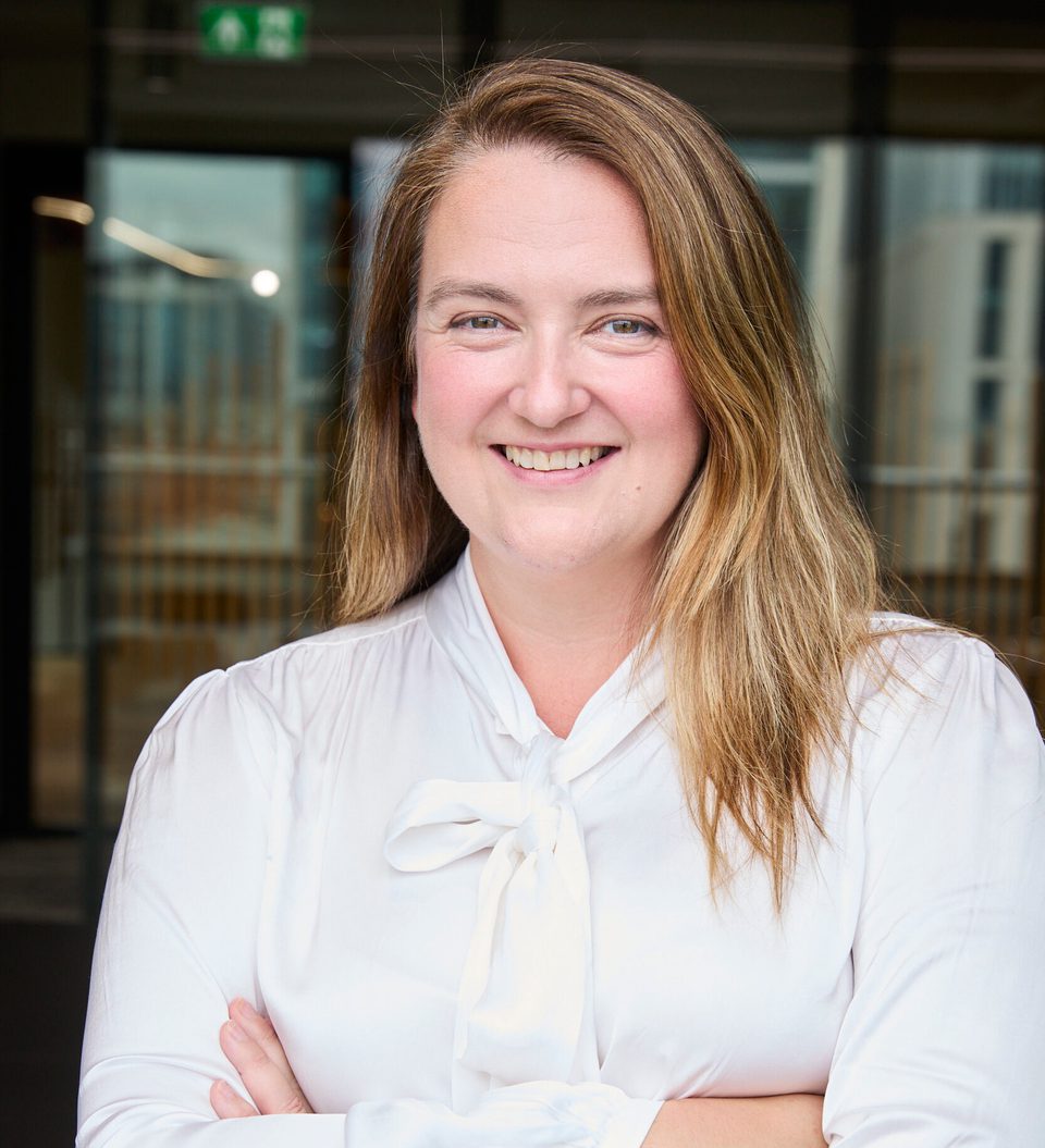 Portrait of a person with long light brown hair, wearing a white blouse with a bow, smiling with arms crossed in a bright office lobby backdrop